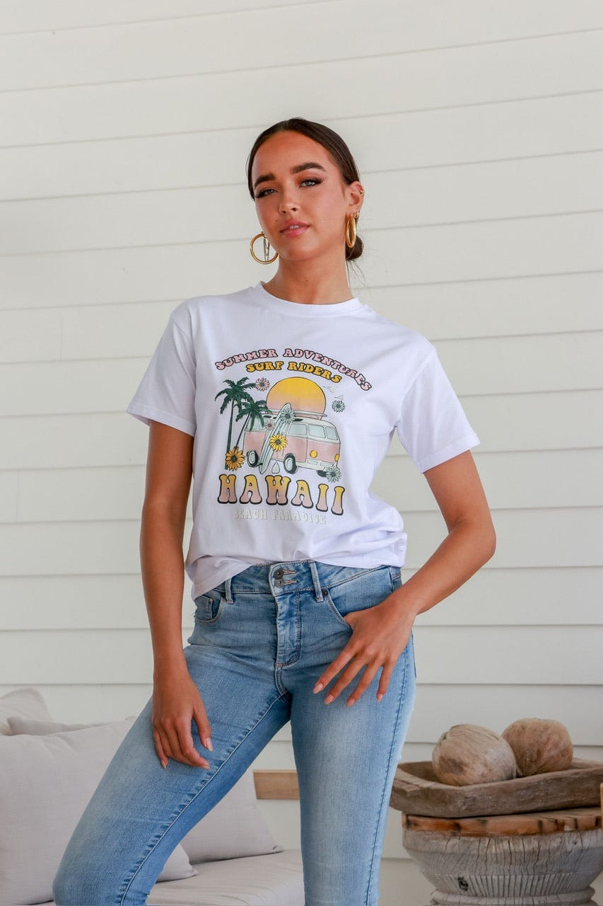 A woman wearing a white crew neck t-shirt with a boho-inspired print, including palm trees and the text 'Hawaii', standing in a casual pose with one hand on her hip. She is also wearing blue jeans.