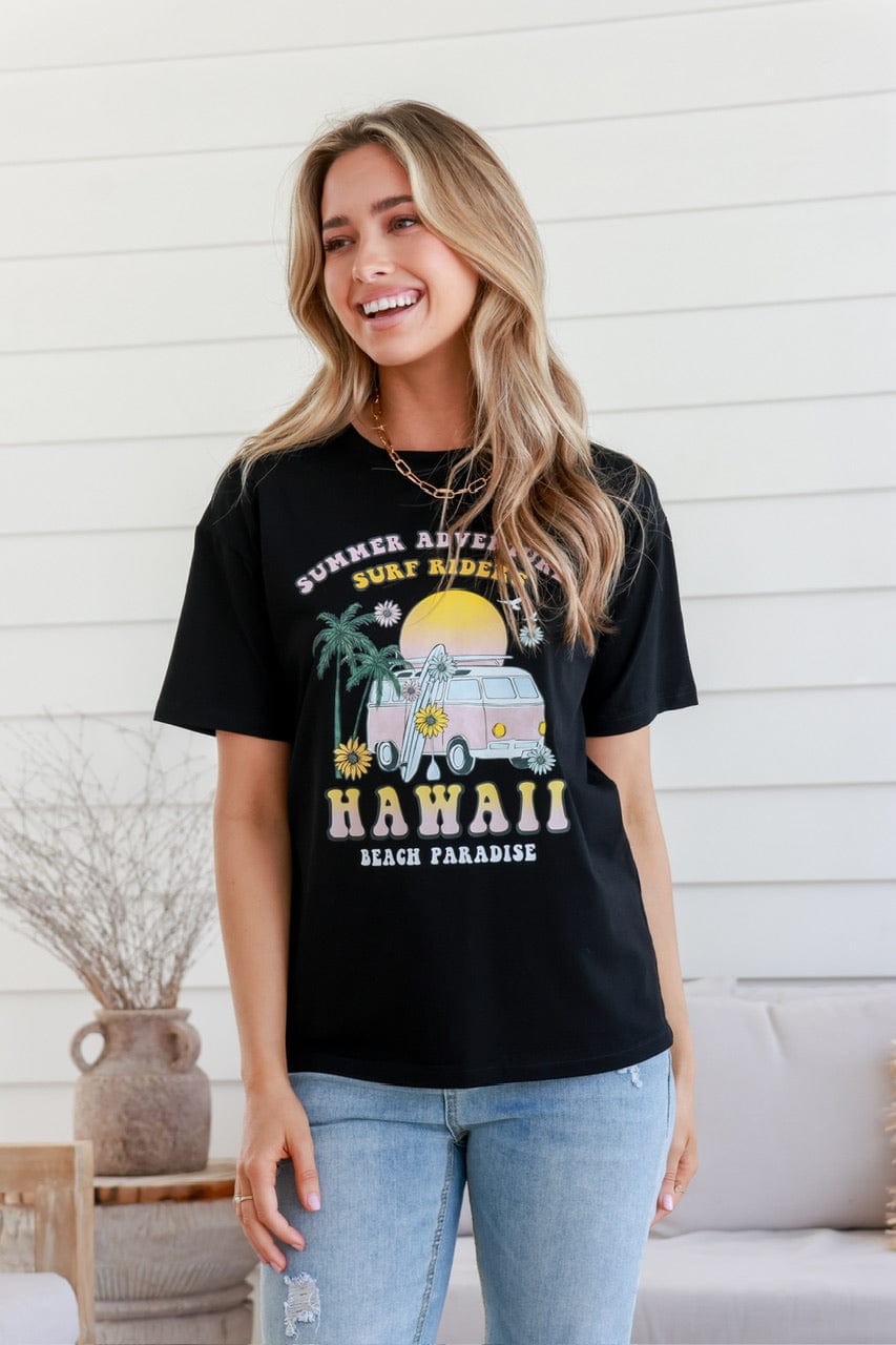 A woman smiling and wearing a black short-sleeved crew neck t-shirt with 'HAWAII' and 'Black Paradise' printed on the front, paired with blue jeans.
