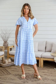 A woman standing in an indoor setting wearing a blue gingham midi dress with short sleeves, a V-neckline, and a tiered design.