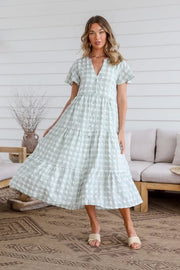 A woman standing in an indoor setting wearing a sage green gingham midi dress with short sleeves, a V-neckline, and a tiered design.
