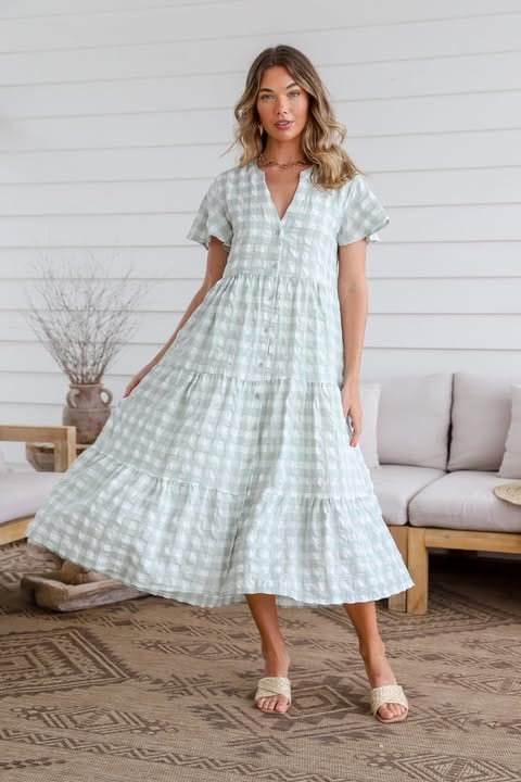 A woman standing in an indoor setting wearing a sage green gingham midi dress with short sleeves, a V-neckline, and a tiered design.