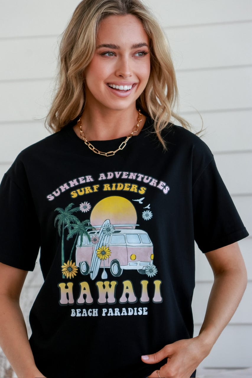 A woman smiling and wearing a black short-sleeved crew neck t-shirt with 'HAWAII' and 'Black Paradise' printed on the front, paired with blue jeans.