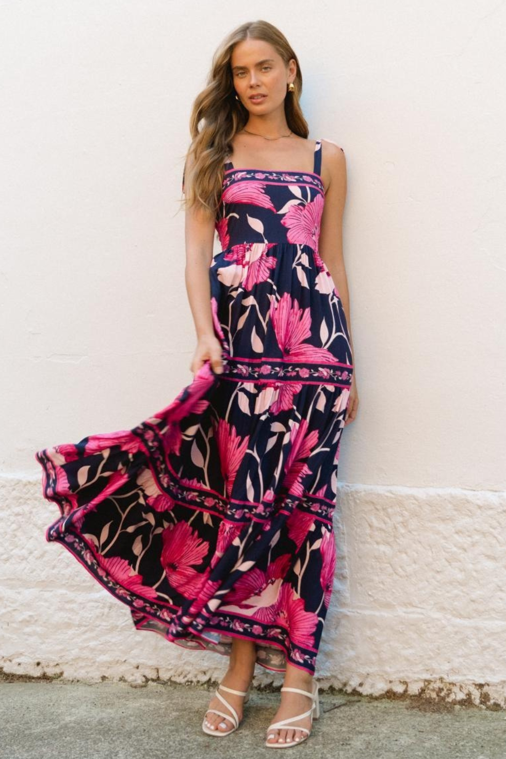 A model wearing a navy and pink floral maxi dress with tie-strap shoulders and a tiered silhouette, standing against a white background.