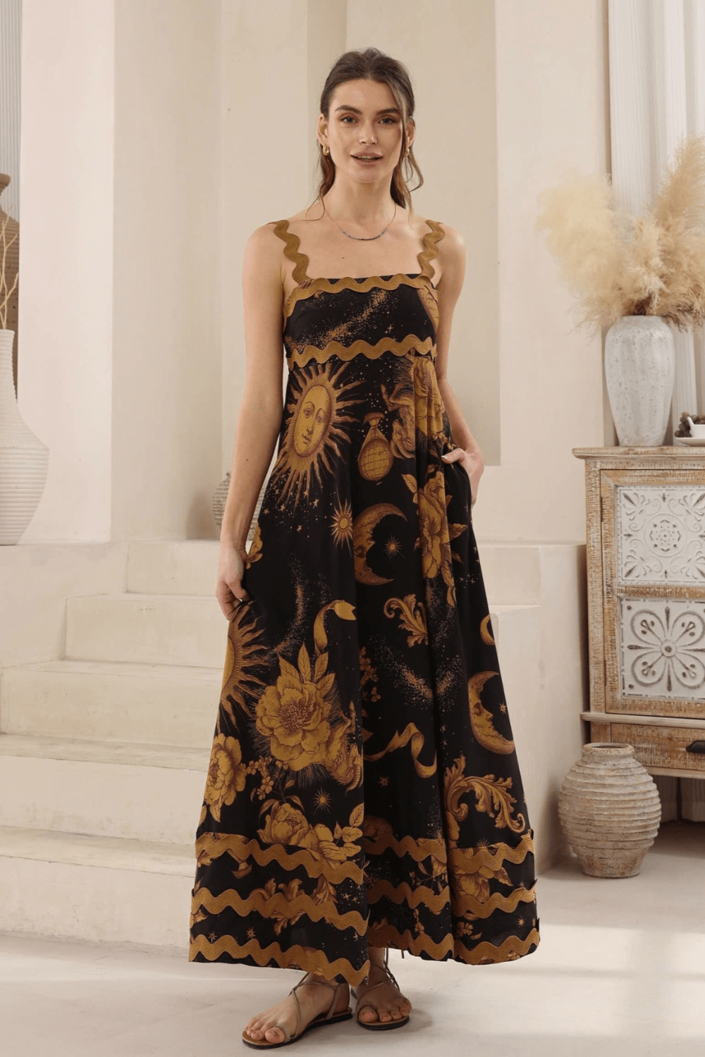 A woman wearing a black maxi dress with a gold Moon and Sun print, and ric rac detailing on the bust and bottom hemline. She is standing in a room with a bohemian style decor.