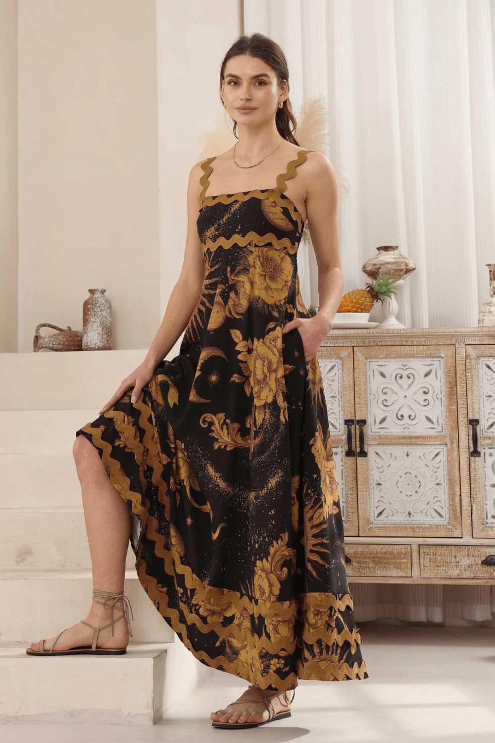 A woman wearing a black maxi dress with a gold Moon and Sun print, and ric rac detailing on the bust and bottom hemline. She is standing in a room with a bohemian style decor.