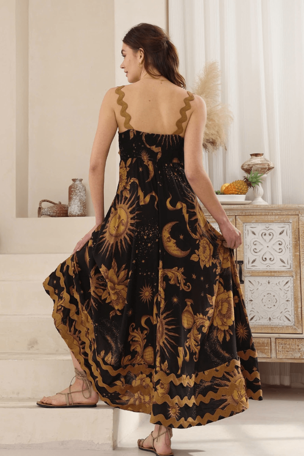 A woman wearing a black maxi dress with a gold Moon and Sun print, and ric rac detailing on the bust and bottom hemline. She is with her back turned standing in a room with a bohemian style decor.