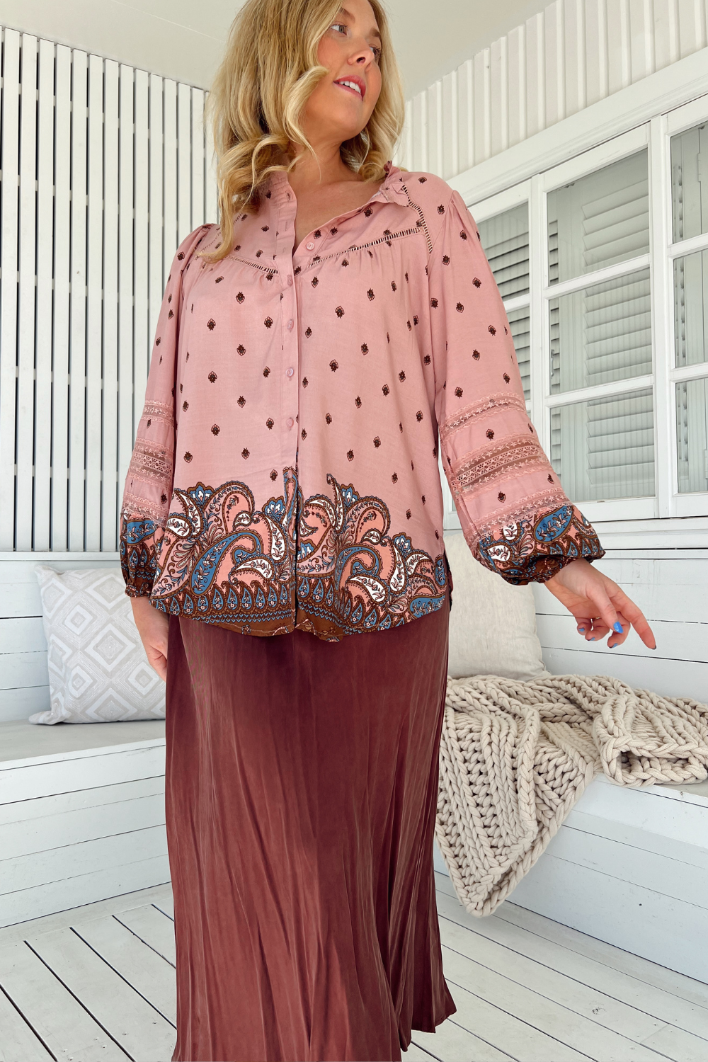 Woman wearing a pink blouse with intricate patterns and a brown skirt in a bright room.