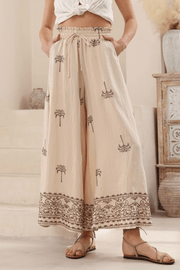 A pair of latte-colored, high-waisted, wide-leg pants with embroidered palm tree motifs and decorative borders, modelled in a casual standing pose.
