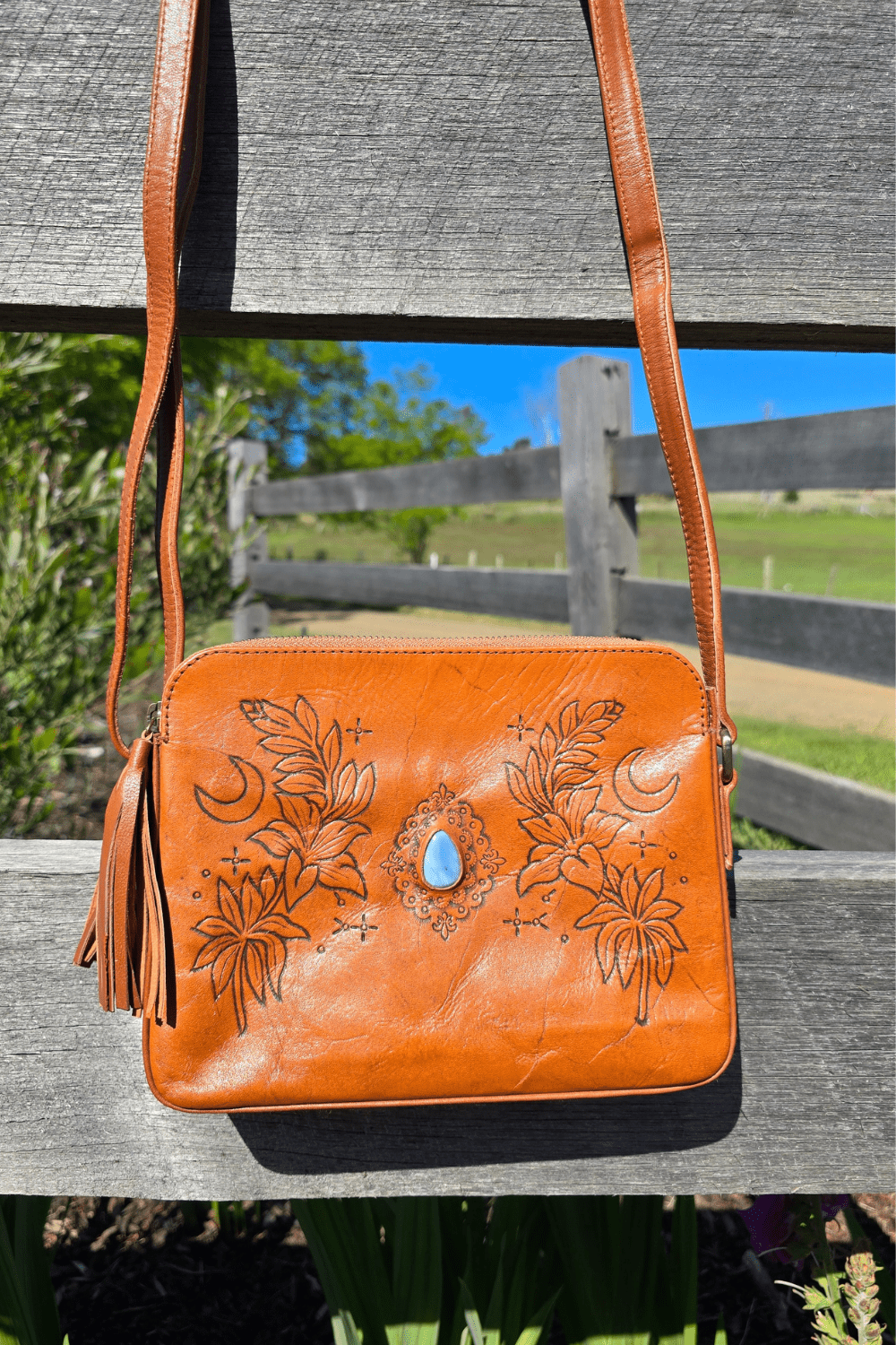 A tan leather shoulder bag with floral embossing and a blue stone, placed on a rock surface with a background of flowers.