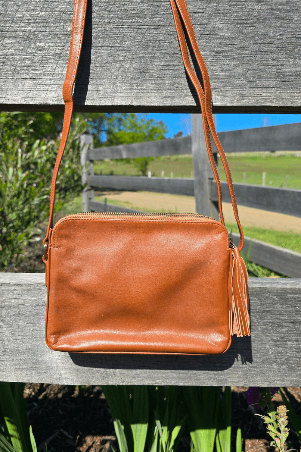 A tan leather shoulder bag with floral embossing and a blue stone, placed on a rock surface with a background of flowers. View of back