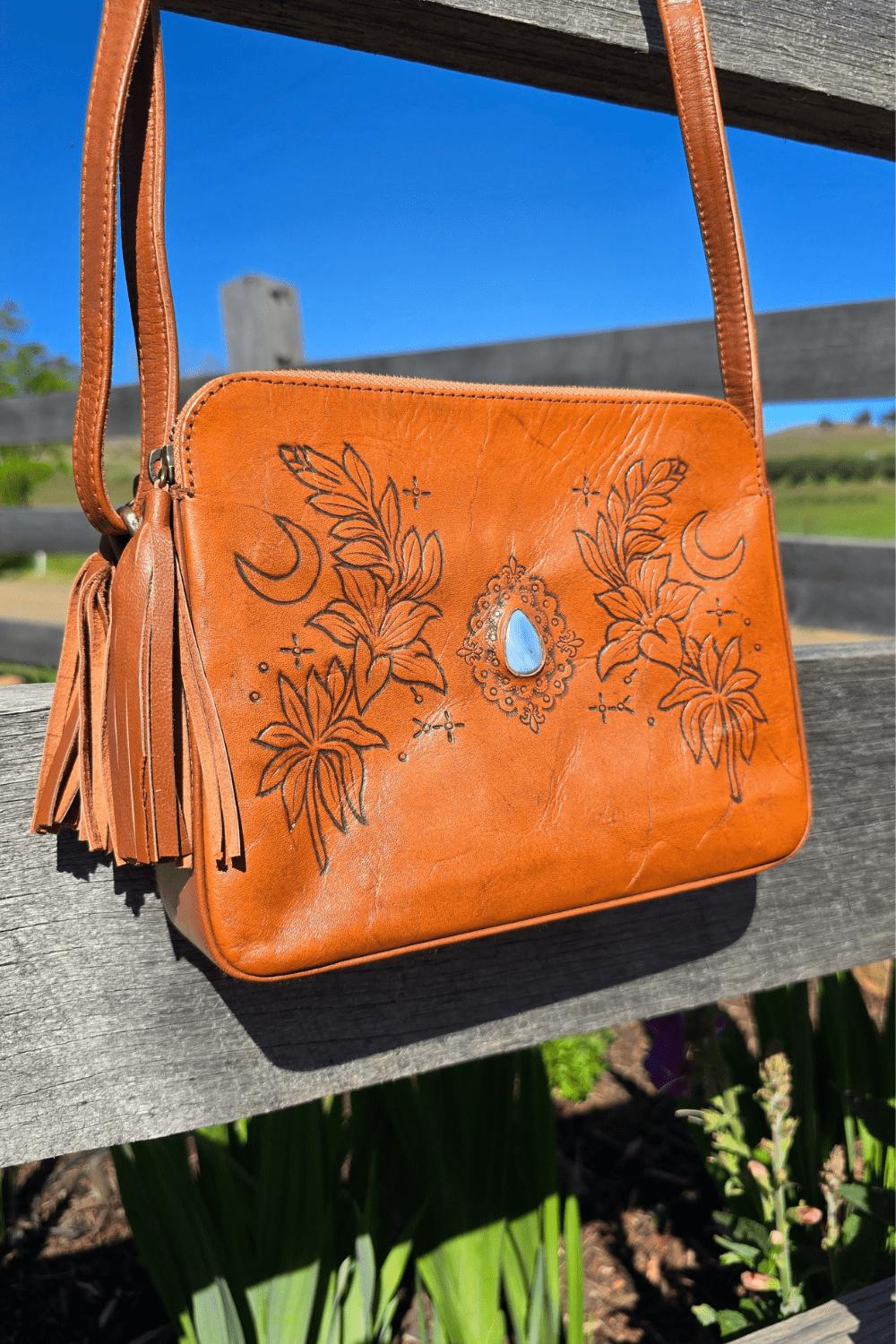 A tan leather shoulder bag with floral embossing and a blue stone, placed on a rock surface with a background of flowers.