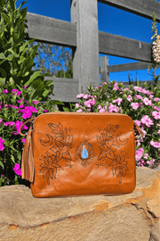 A tan leather shoulder bag with floral embossing and a blue stone, placed on a rock surface with a background of flowers.