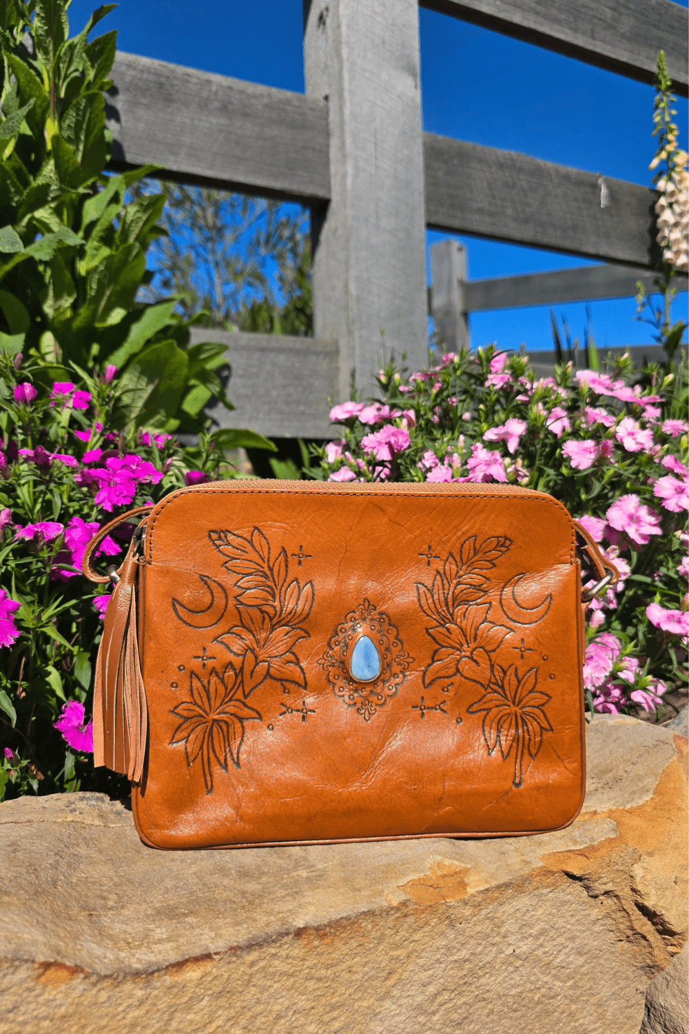A tan leather shoulder bag with floral embossing and a blue stone, placed on a rock surface with a background of flowers.