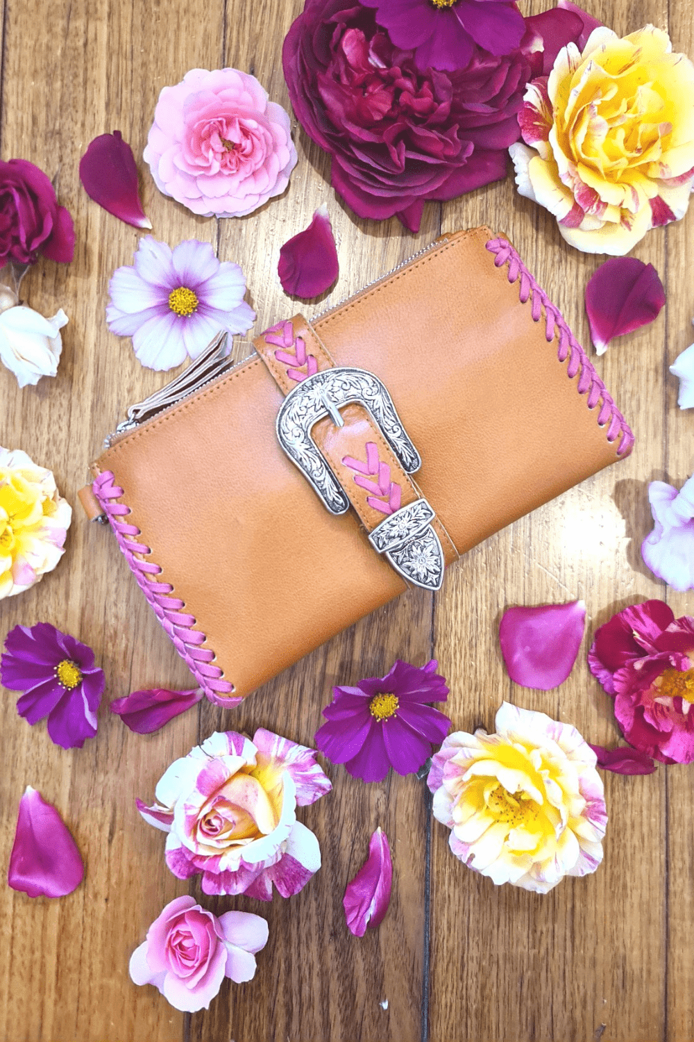 A bohemian leather wallet with a tan base and pink detailing, laid out on a surface surrounded by various flowers.