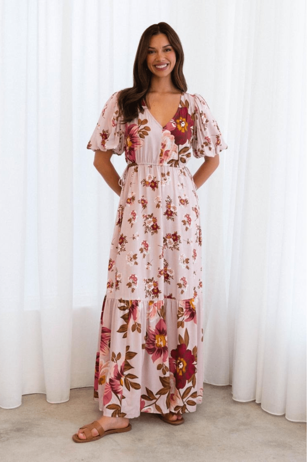 A woman wearing a floral maxi dress with a flattering v-neckline, short balloon sleeves with elastic cuffs, and an adjustable drawstring waist with side ties.