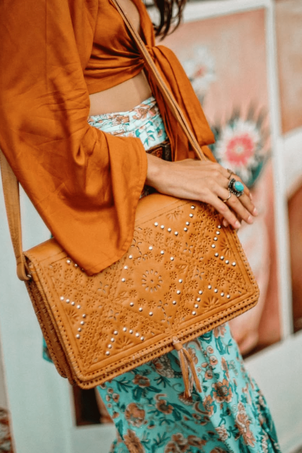 A person holding a tan leather satchel handbag with intricate hand-tooled details and stud embellishments.