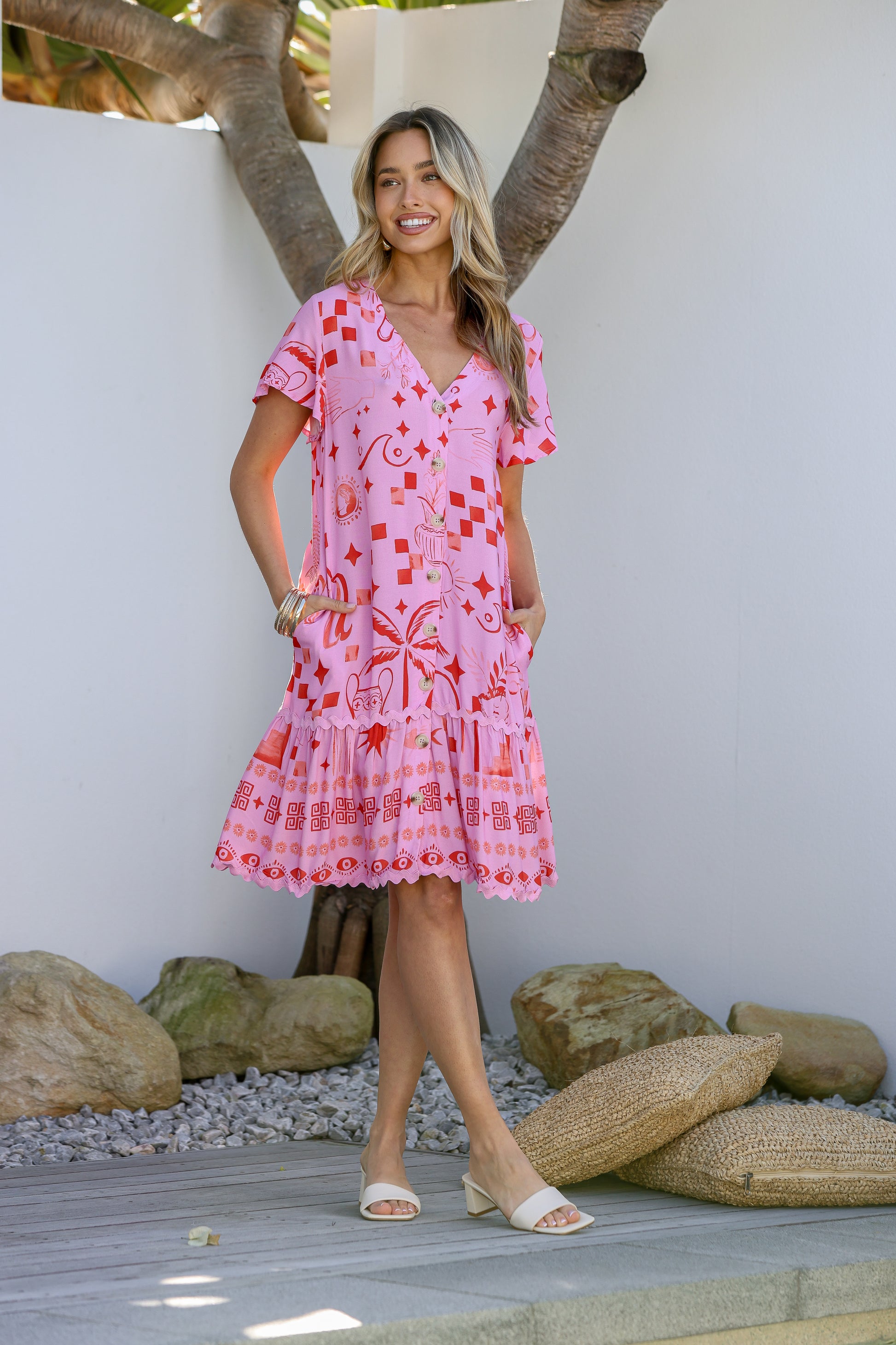 A woman wearing a short, pink dress with a red floral pattern and ric rac trim, standing near a tree and rocks.