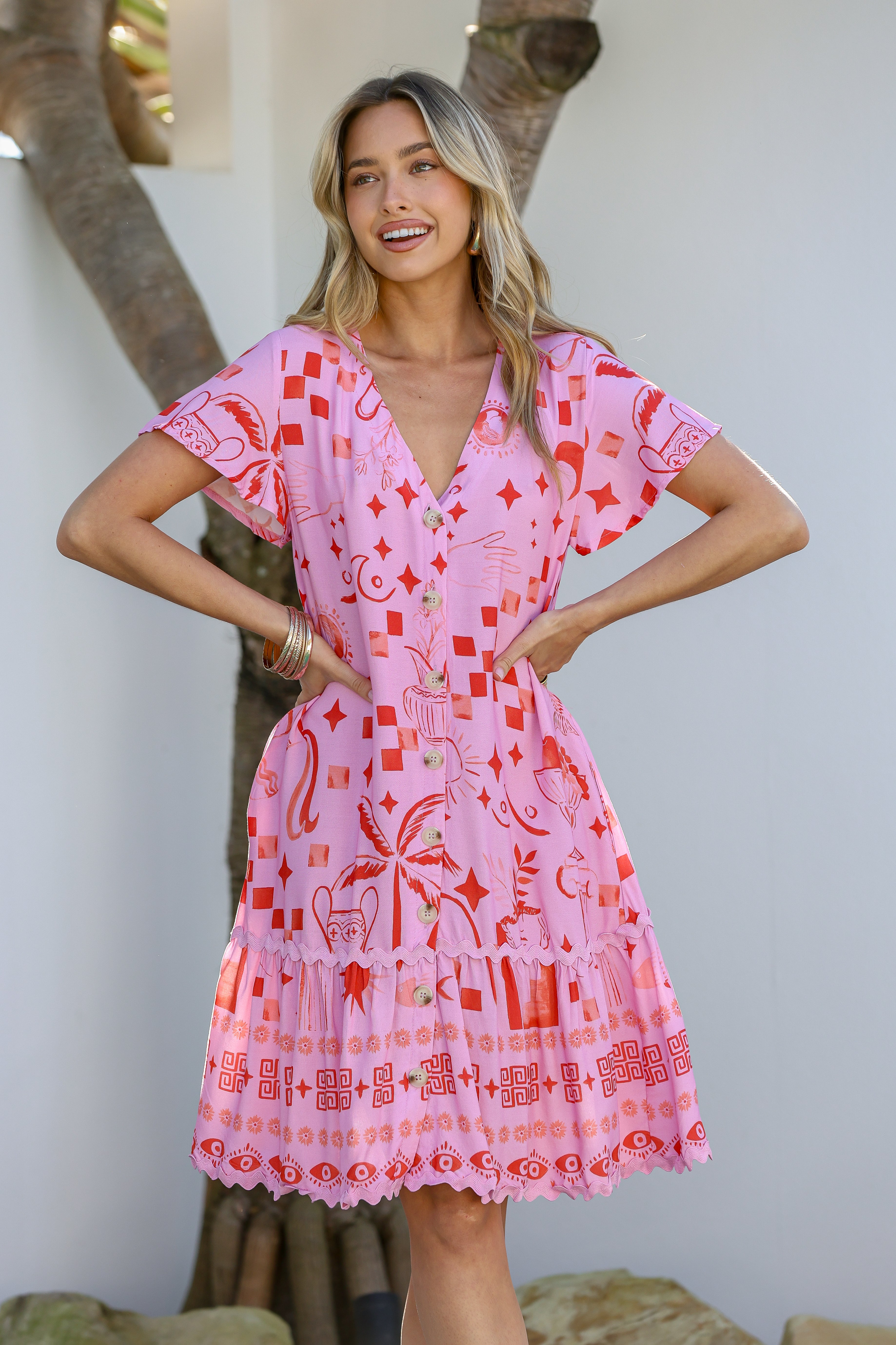 A woman wearing a pink mini dress with a tropical-inspired print and ric rac trim, featuring a V-neckline and short sleeves.