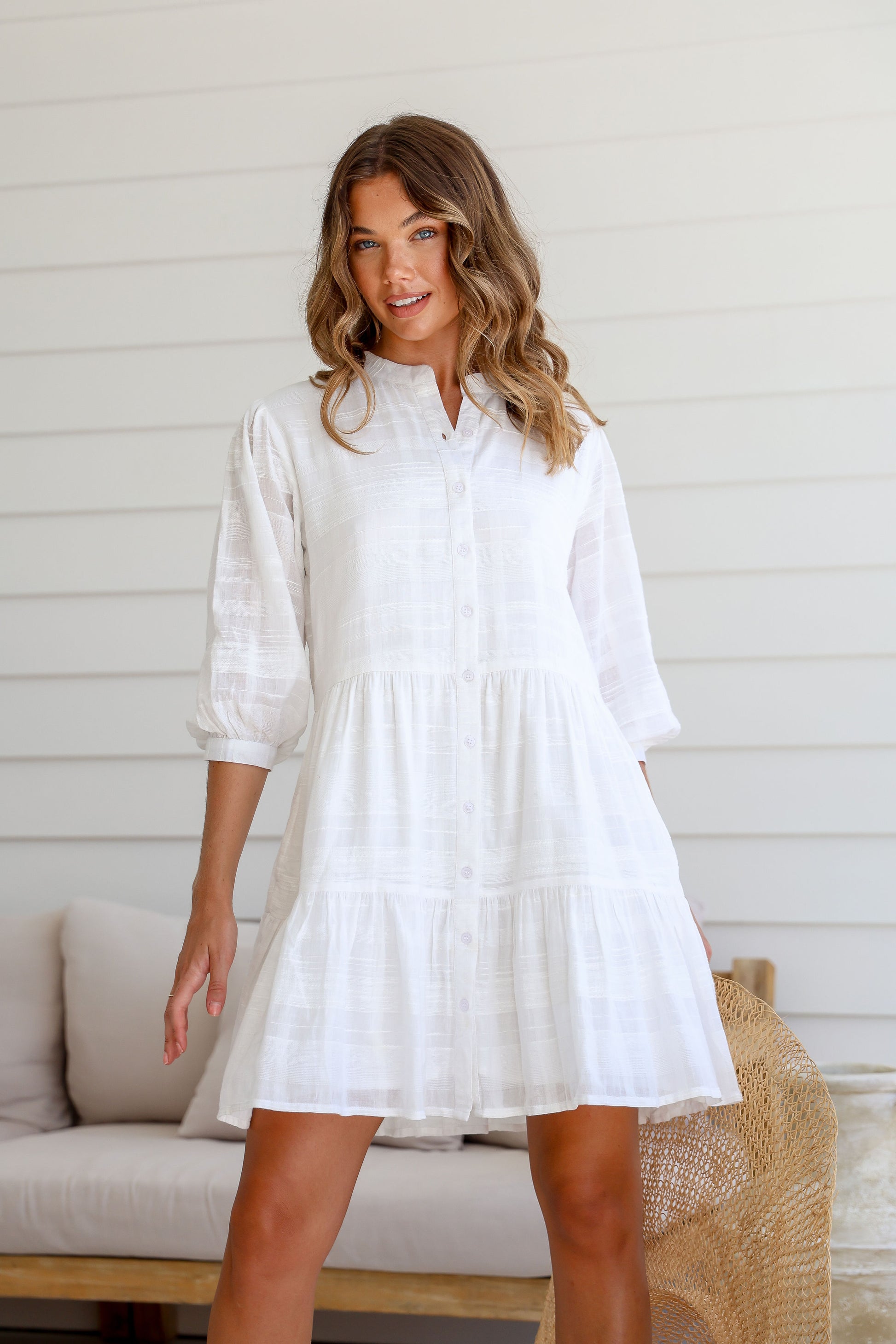 A woman standing in a casual setting wearing a white cotton mini dress with a collared neckline, 3/4 sleeves with soft gathers, and a tiered skirt silhouette. The dress features functional side pockets and a button-through front.