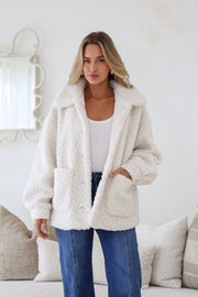 A woman wearing a white teddy boucle jacket with an oversized shawl collar, full button-through front, and two roomy front pockets. She is also wearing blue jeans and has her hands in the pockets.