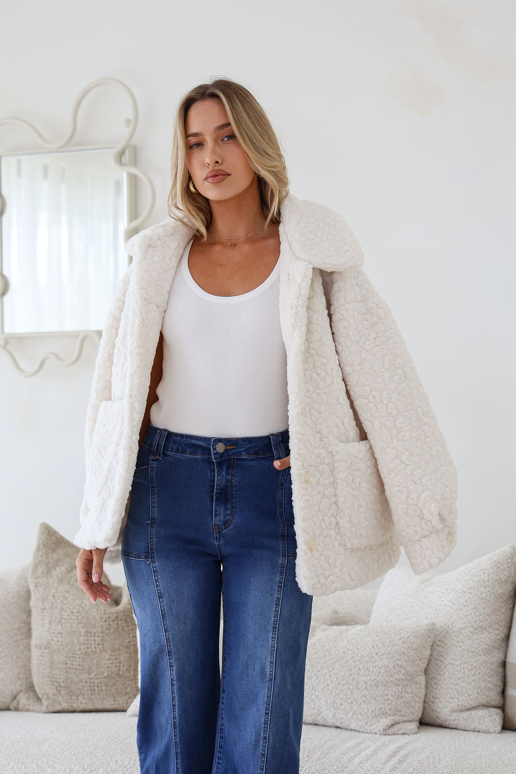 Woman wearing a white fluffy coat, white top, and blue jeans in a room with a mirror and pillows.