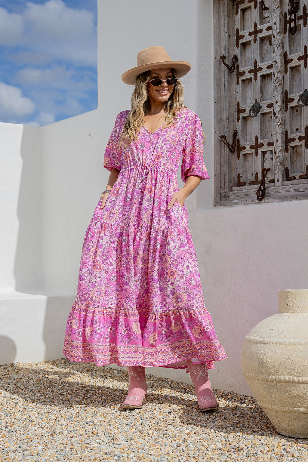 Woman in a pink floral dress and hat standing in front of a white wall with a textured door.