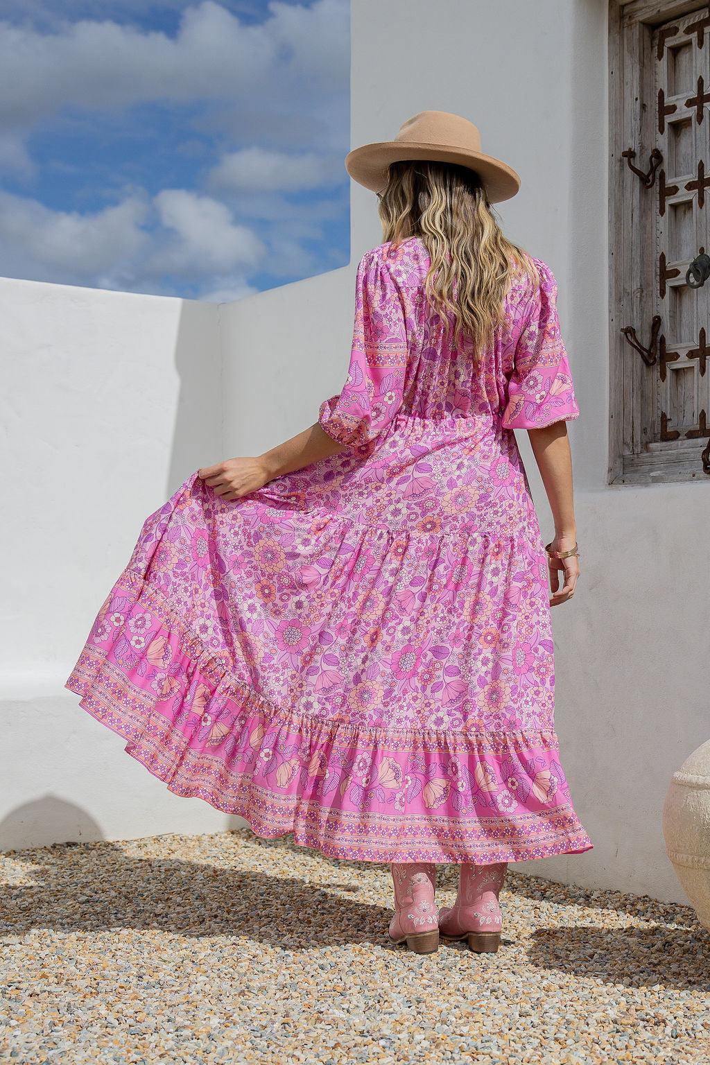 Woman in a pink dress and hat standing against a white wall with blue sky.