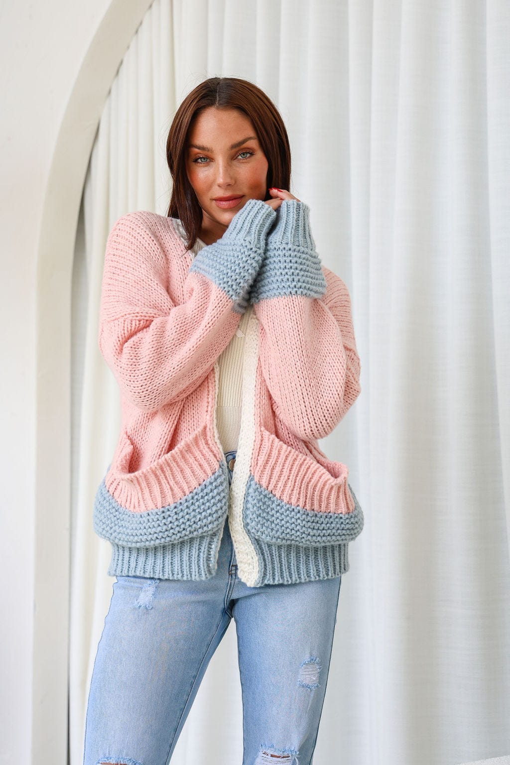 A woman wearing a chunky knit cardigan in pink and blue colors, with long sleeves and an open front, paired with ripped jeans.