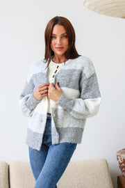 A woman wearing a grey striped cardigan with chunky buttons over a white top, paired with blue jeans and standing in a room with a neutral decor.