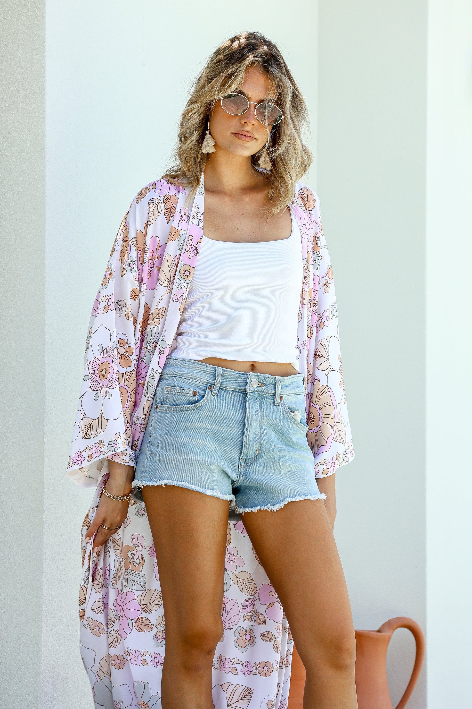 A woman standing outdoors wearing a floral kimono with boho-inspired border, paired with a white tank top and blue shorts.