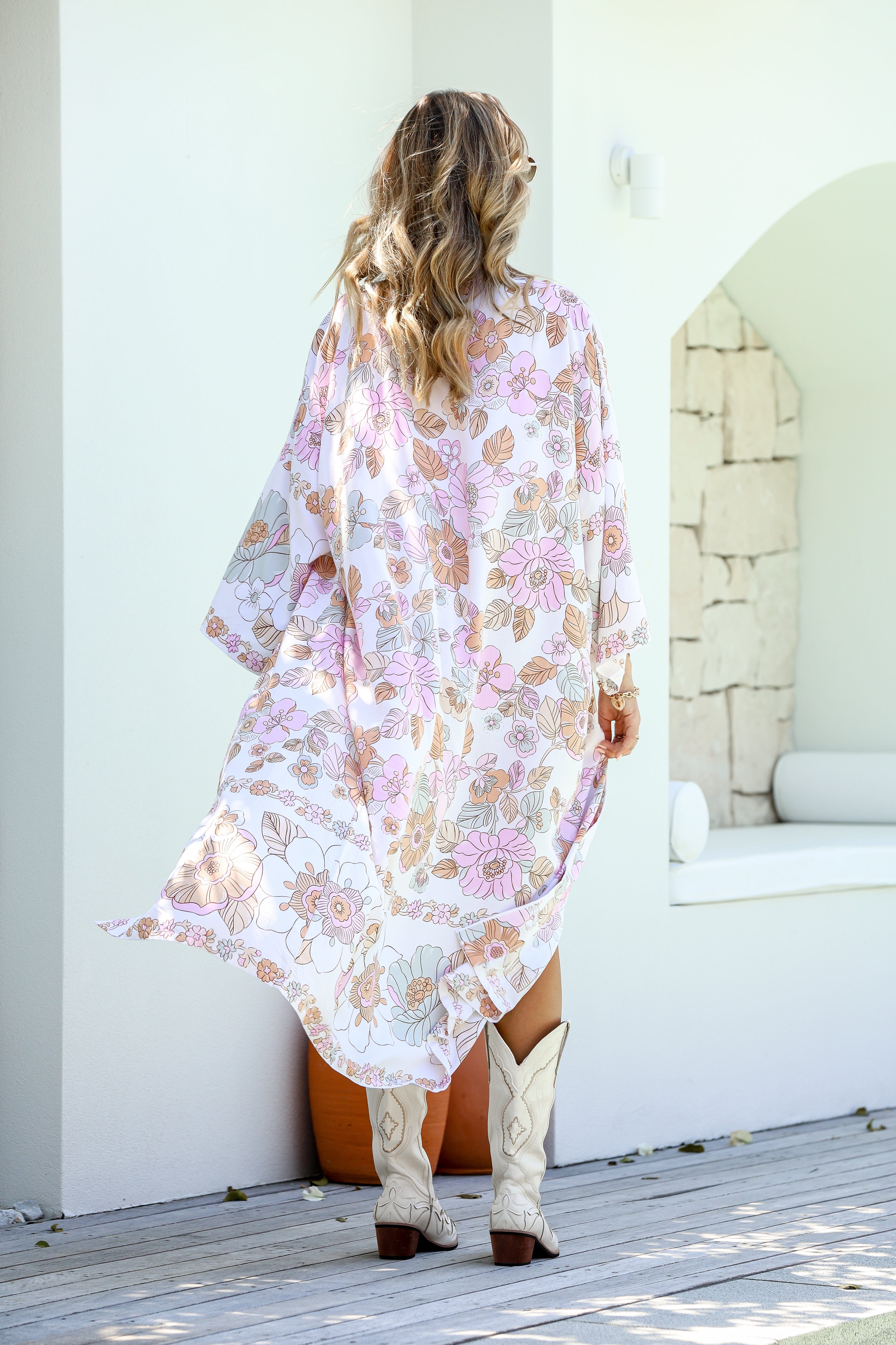 A woman standing outdoors wearing a floral kimono with boho-inspired border, paired with a white tank top and blue shorts.