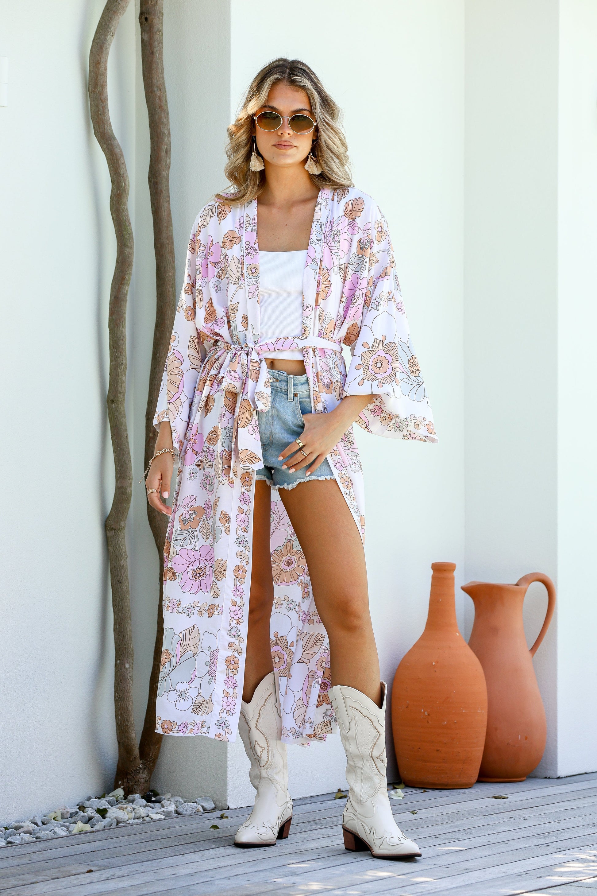 A woman standing outdoors wearing a floral kimono with boho-inspired border, paired with a white tank top and blue shorts.