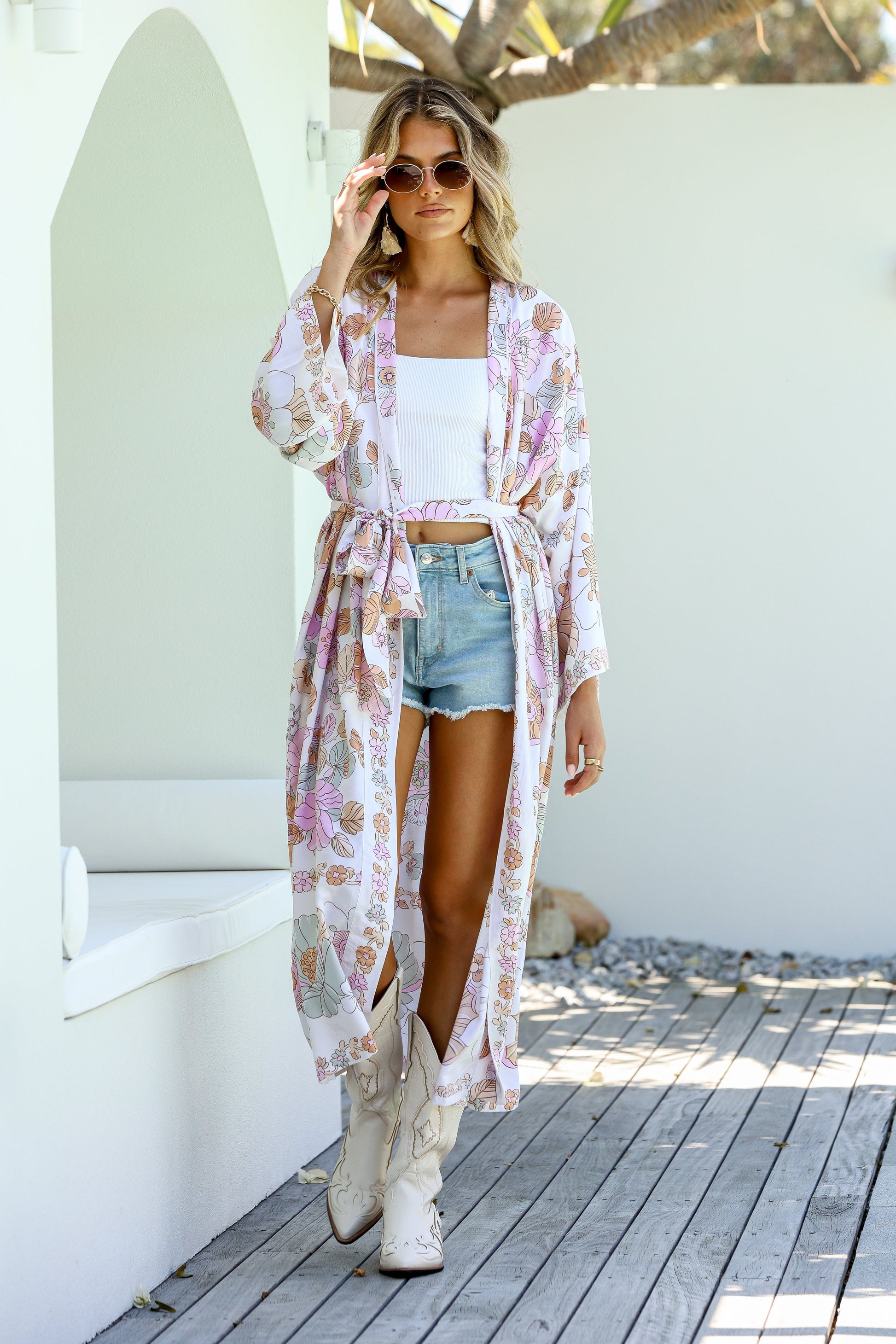 A woman standing outdoors wearing a floral kimono with boho-inspired border, paired with a white tank top and blue shorts.