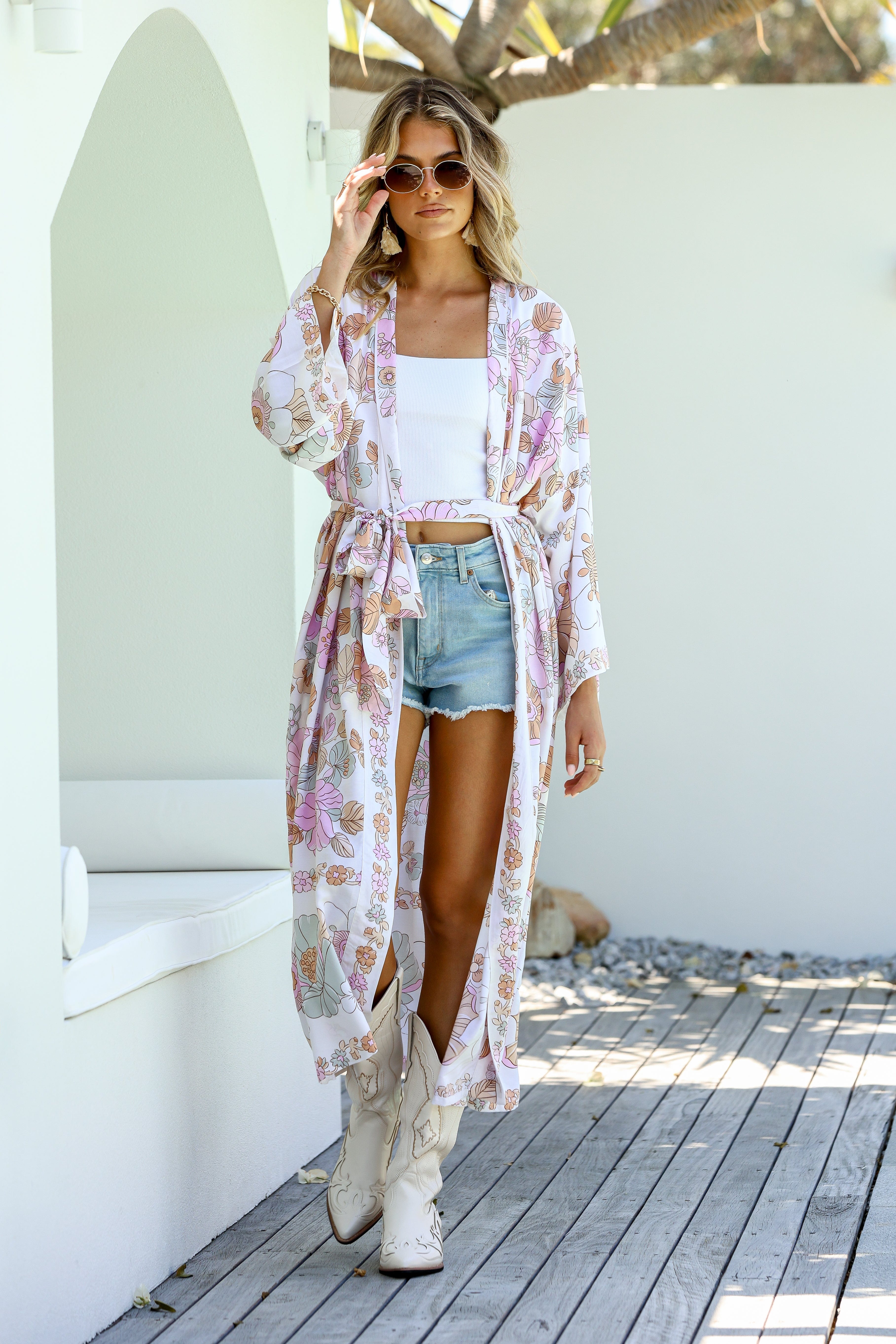 A woman standing outdoors wearing a floral kimono with boho-inspired border, paired with a white tank top and blue shorts.