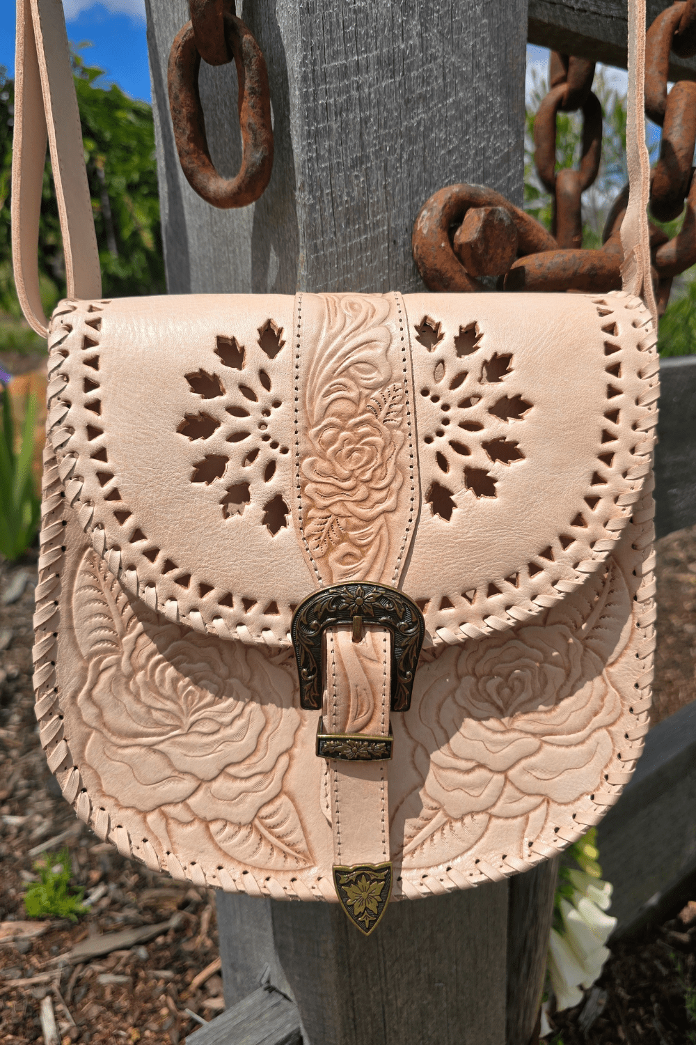 A natural-colored leather crossbody bag with floral hand-tooled detailing, an antiqued brass-style buckle, and an adjustable strap, displayed against a rustic backdrop with a wooden fence and flowers.