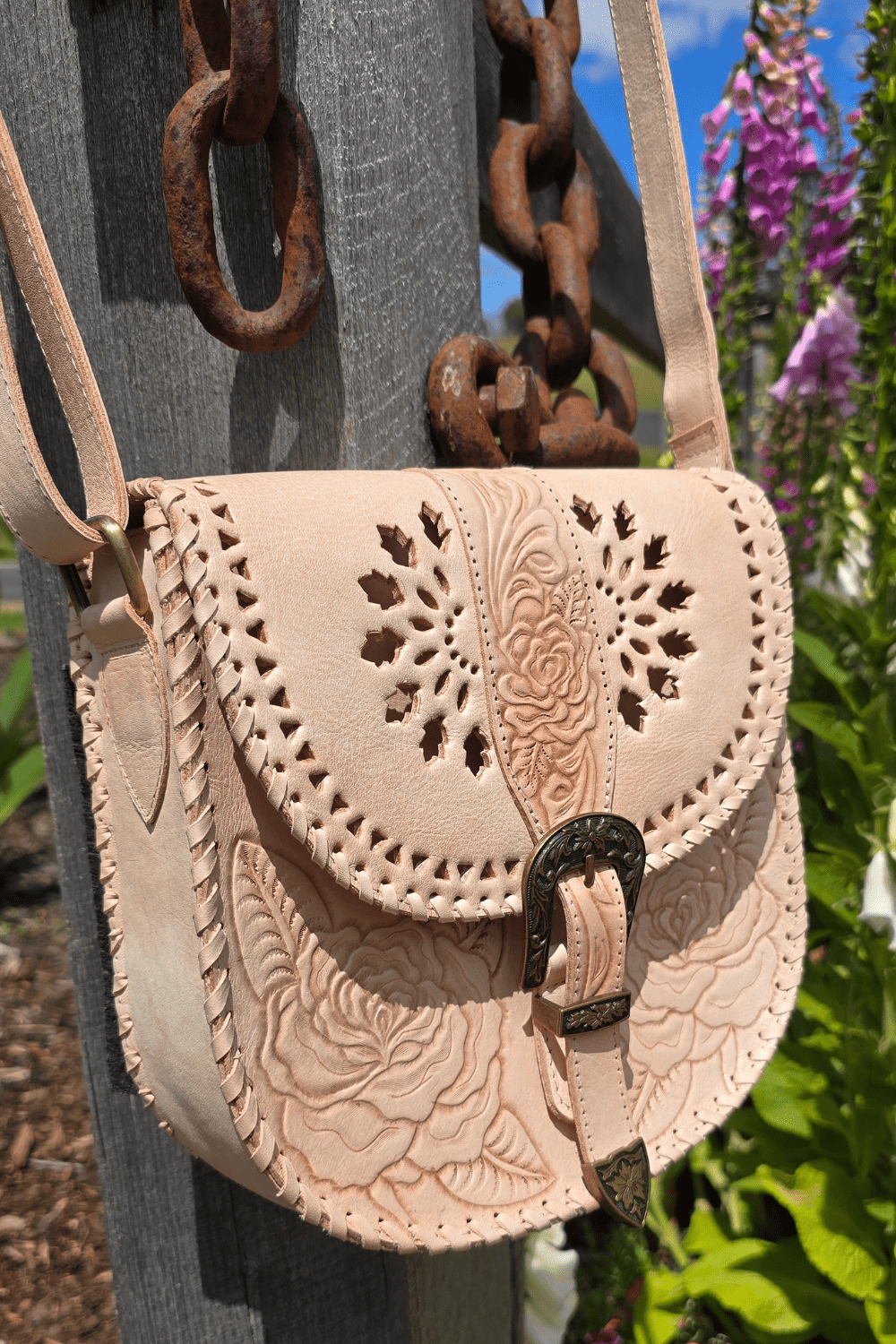 A natural-colored leather crossbody bag with floral hand-tooled detailing, an antiqued brass-style buckle, and an adjustable strap, displayed against a rustic backdrop with a wooden fence and flowers.