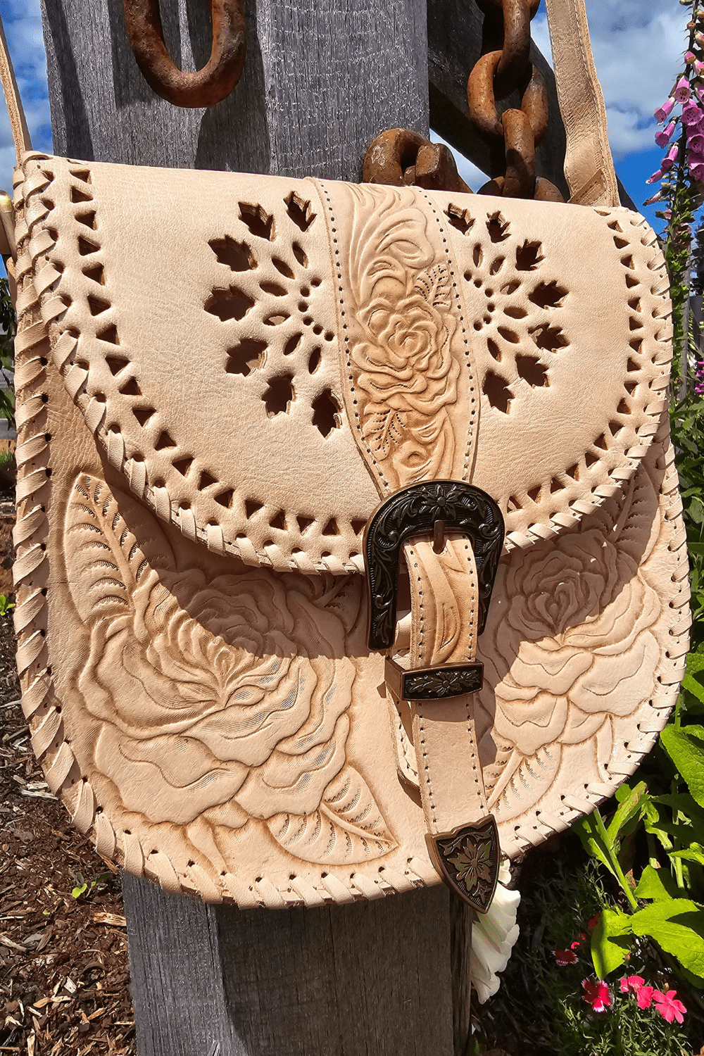A natural-colored leather crossbody bag with floral hand-tooled detailing, an antiqued brass-style buckle, and an adjustable strap, displayed against a rustic backdrop with a wooden fence and flowers.