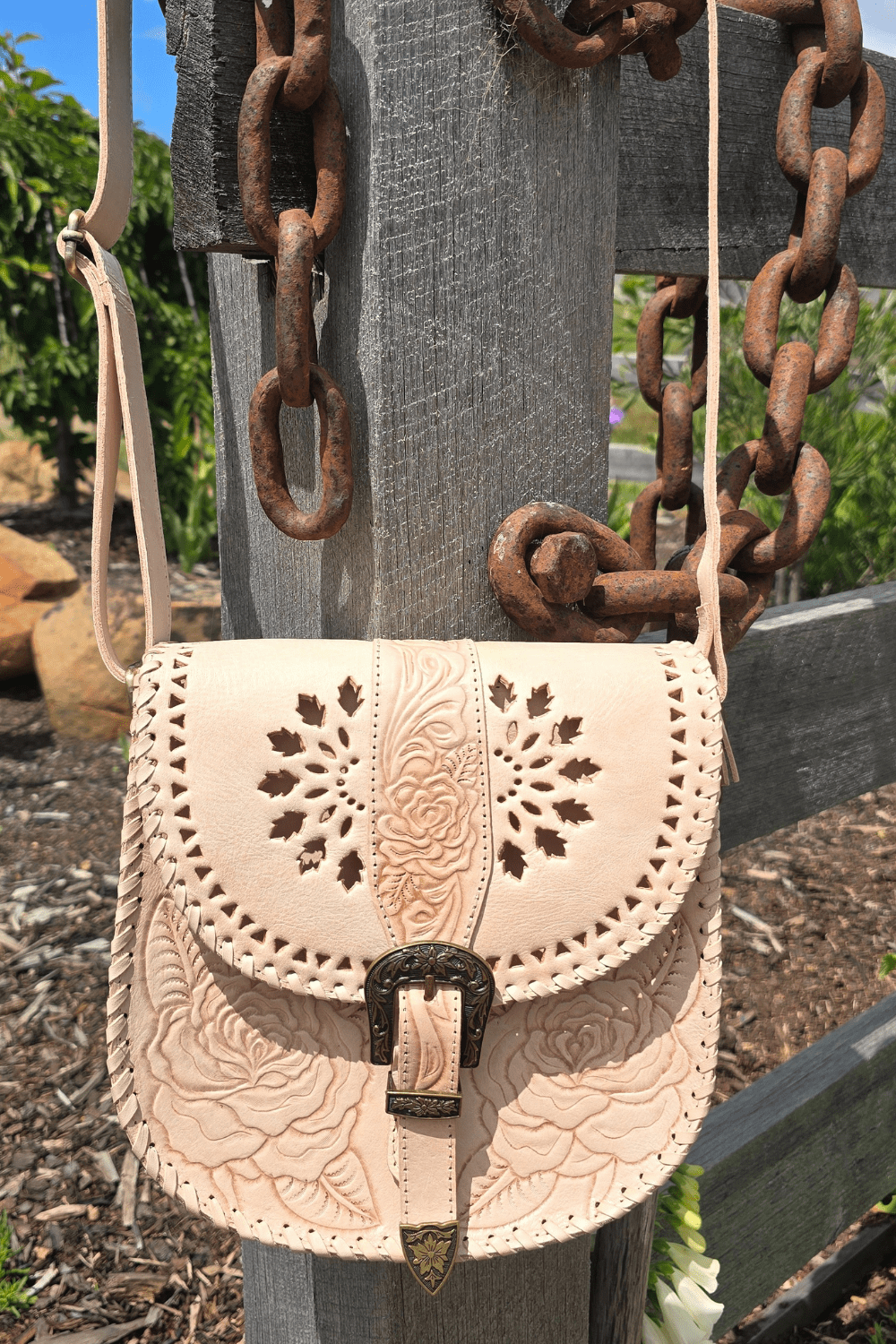 A natural-colored leather crossbody bag with floral hand-tooled detailing, an antiqued brass-style buckle, and an adjustable strap, displayed against a rustic backdrop with a wooden fence and flowers.