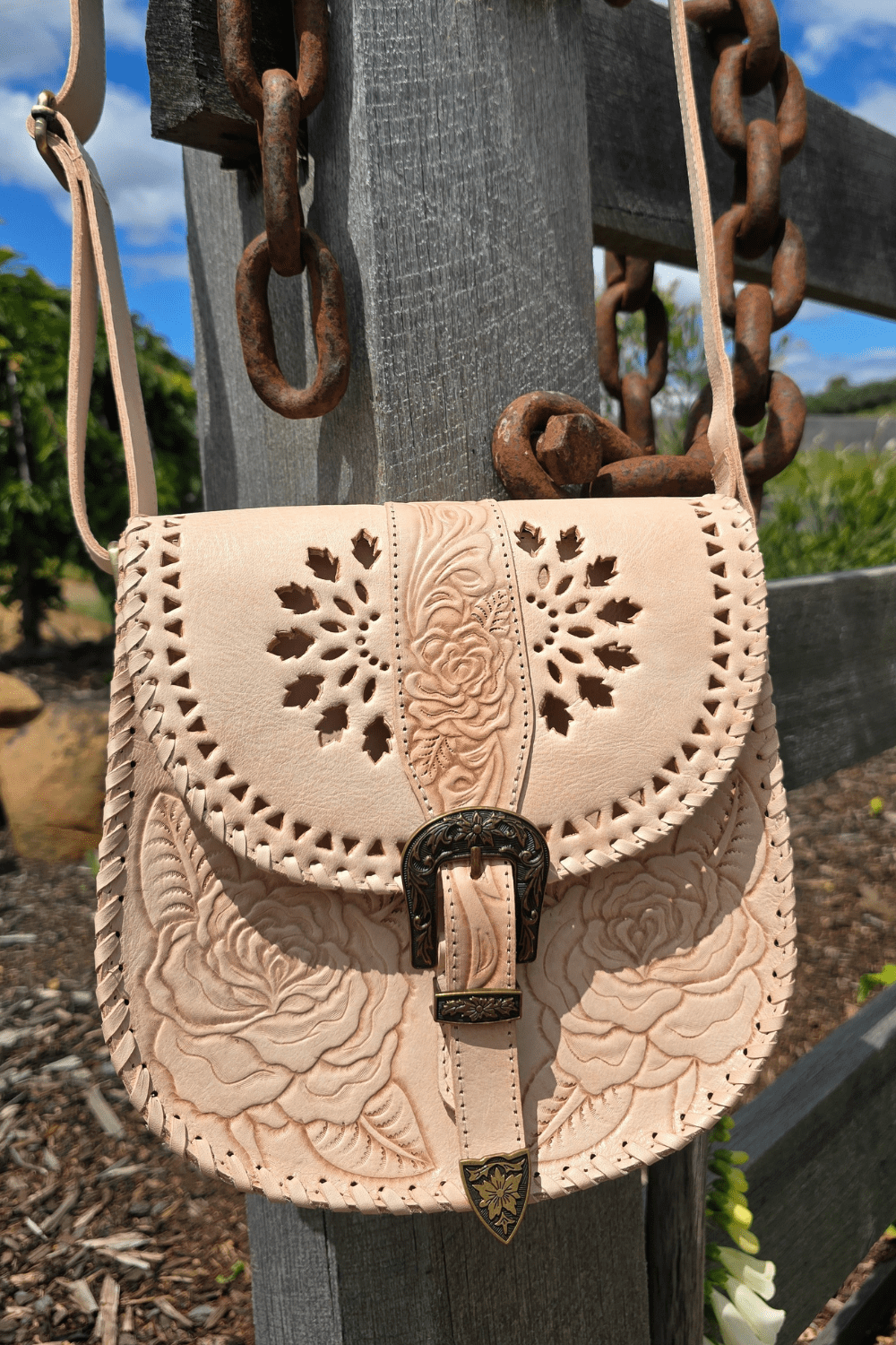 A natural-colored leather crossbody bag with floral hand-tooled detailing, an antiqued brass-style buckle, and an adjustable strap, displayed against a rustic backdrop with a wooden fence and flowers.