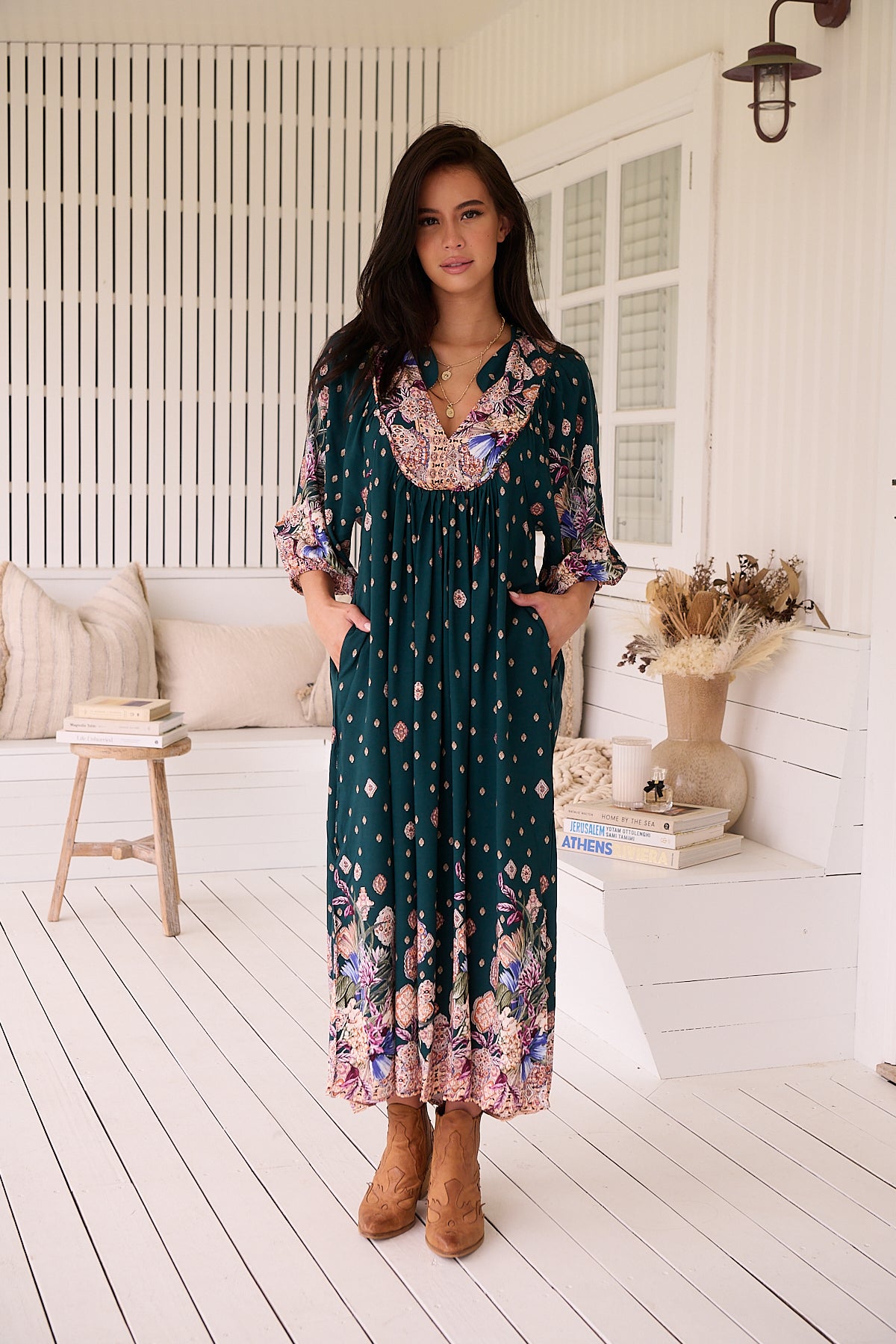 A woman standing in a room with white panelled walls, wearing a floral bohemian midi dress with a dark green base and long sleeves.