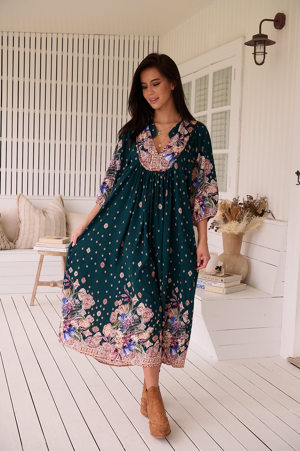 A woman standing in a room with white panelled walls, wearing a floral bohemian midi dress with a dark green base and long sleeves.