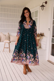 A woman standing in a room with white panelled walls, wearing a floral bohemian midi dress with a dark green base and long sleeves.