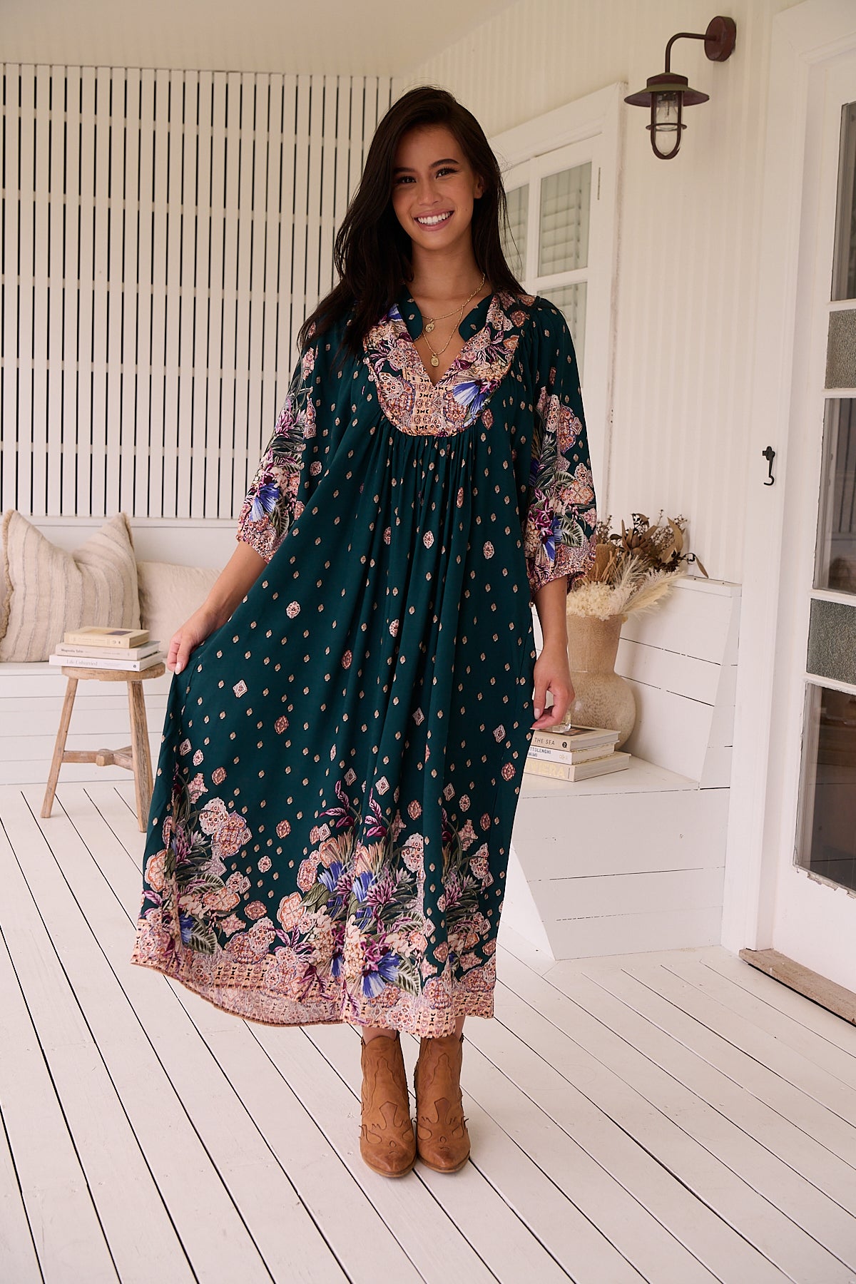 A woman standing in a room with white panelled walls, wearing a floral bohemian midi dress with a dark green base and long sleeves.