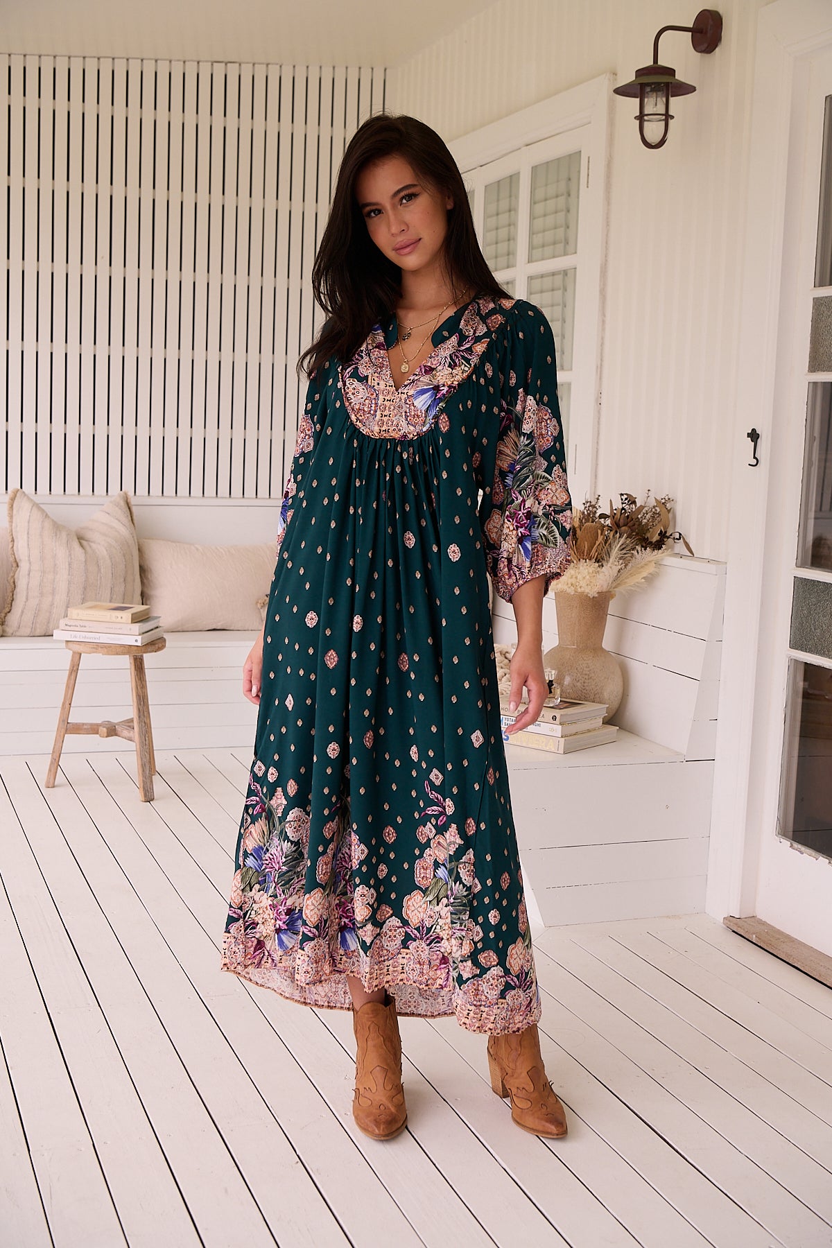 A woman standing in a room with white panelled walls, wearing a floral bohemian midi dress with a dark green base and long sleeves.