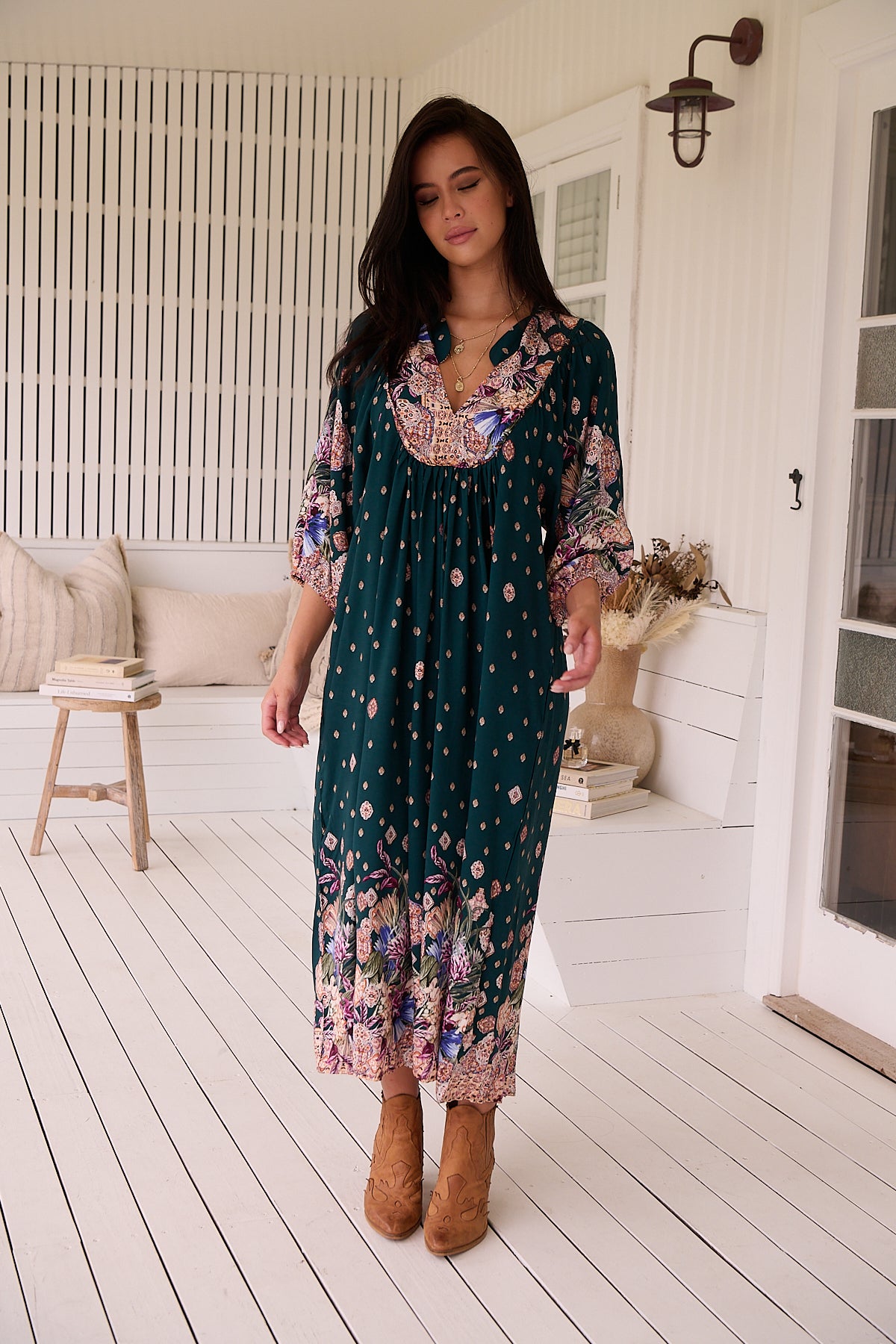 A woman standing in a room with white panelled walls, wearing a floral bohemian midi dress with a dark green base and long sleeves.
