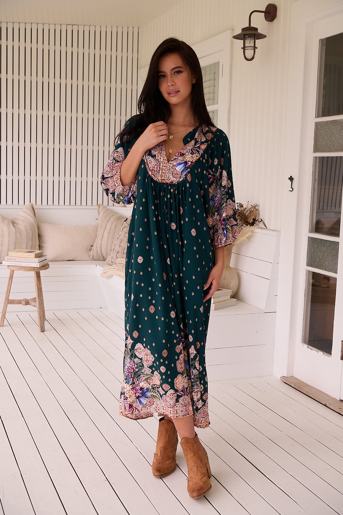 A woman standing in a room with white panelled walls, wearing a floral bohemian midi dress with a dark green base and long sleeves.