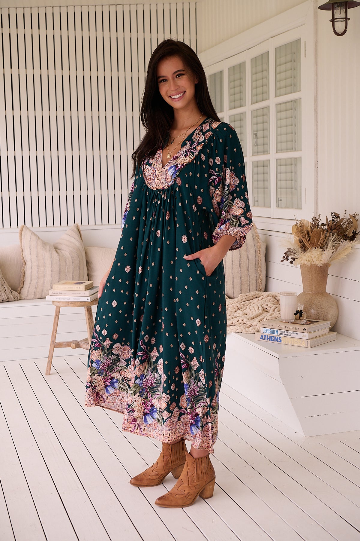 A woman standing in a room with white panelled walls, wearing a floral bohemian midi dress with a dark green base and long sleeves.