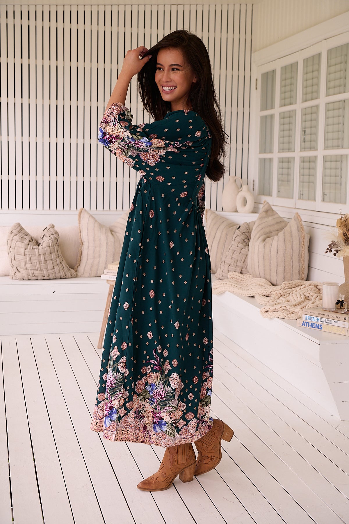 A woman standing in a room with white panelled walls, wearing a floral bohemian midi dress with a dark green base and long sleeves.