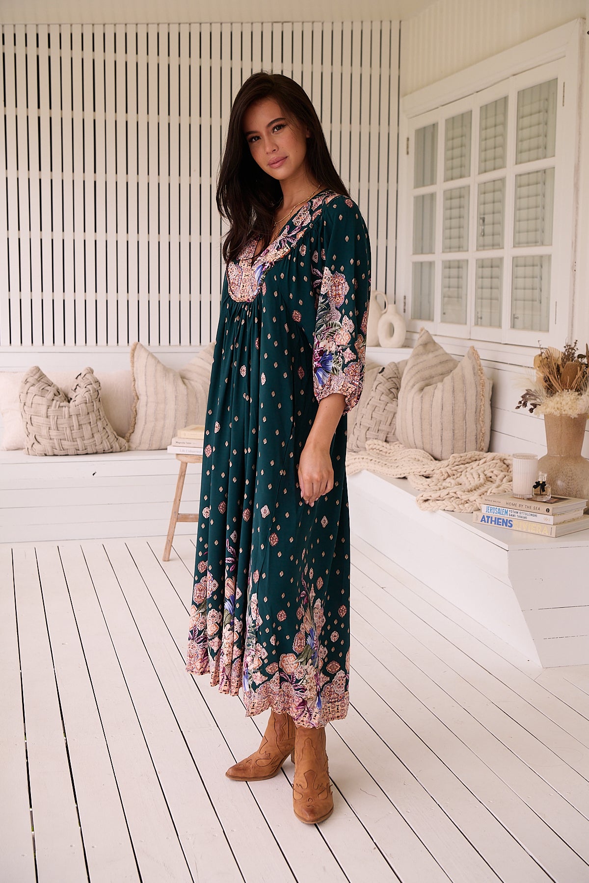 A woman standing in a room with white panelled walls, wearing a floral bohemian midi dress with a dark green base and long sleeves.