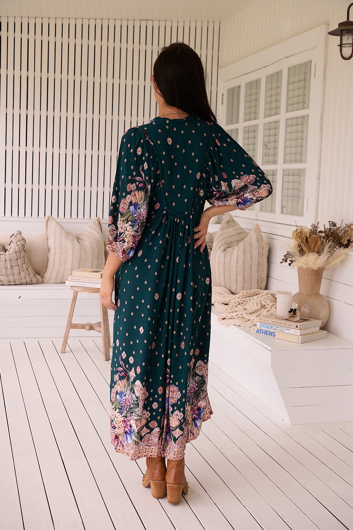 A woman standing in a room with white panelled walls, wearing a floral bohemian midi dress with a dark green base and long sleeves.